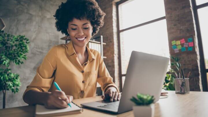 Mulher sentada com computador aberto e escrevendo em um caderno