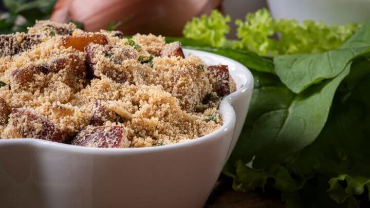 Recipiente branco com farofa e bacon em cima de uma mesa com cebola e folhas verdes