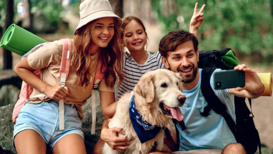 "Pai, mãe e filha, com cachorro Golden Retriever, tirando selfie com mochilas nas costas, indicando viagem