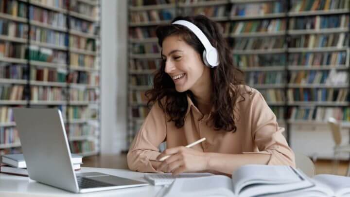 Mulher usando fones de ouvido sentada de frente para o computador estudando e com livros abertos na mesa