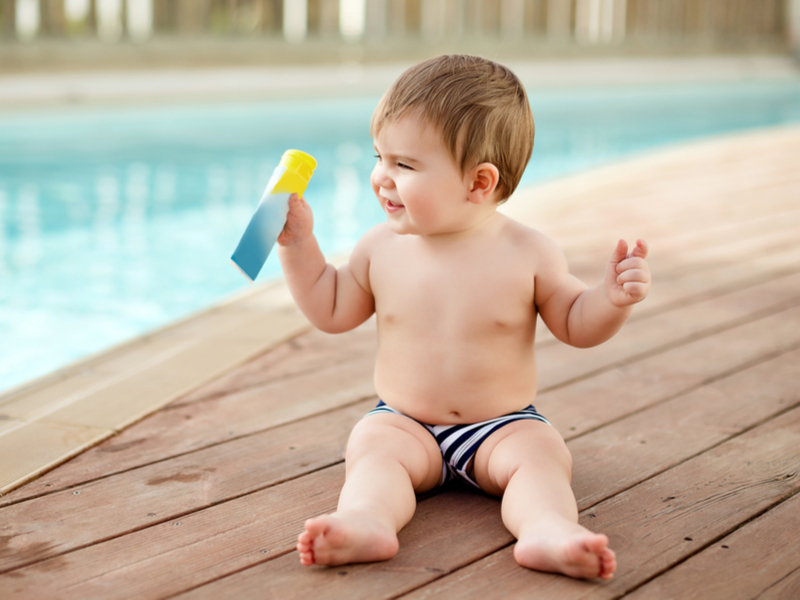 Bebê sentado na beira da piscina segurando um tubo de protetor solar