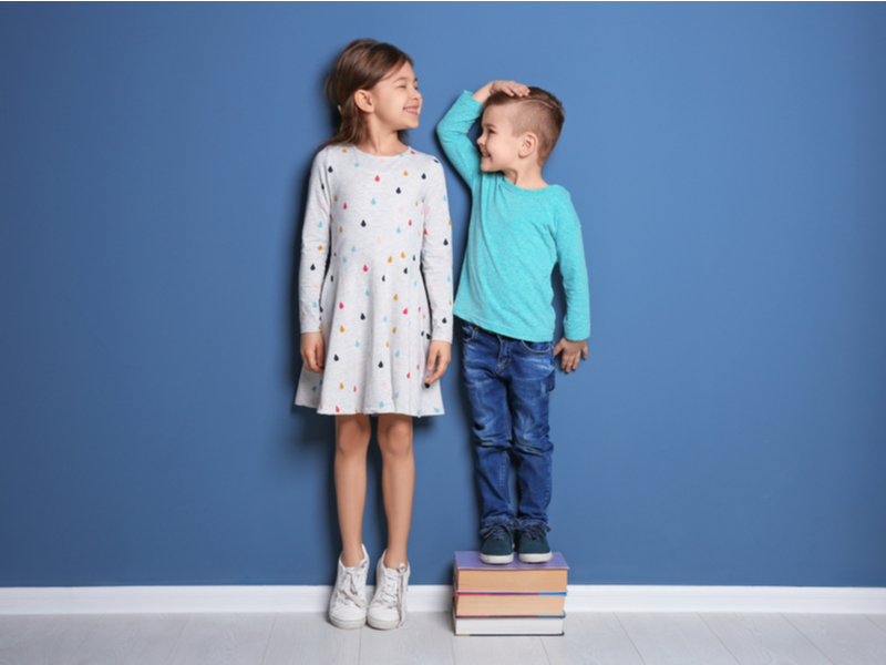 Menino e menina comparando altura em parede azul. Menino é menor e está em cima de pilha de livros
