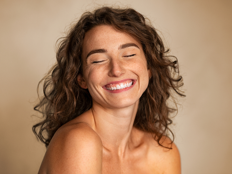 Mulher de cabelo cacheado, sardas na rosto, olhos fechados e sorrindo