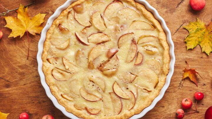 Torta de maçã em recipiente branco, sobre superfície marrom com frutas vermelhas e folhas em volta