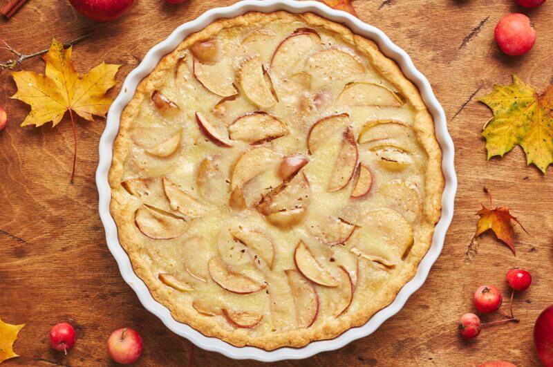 Torta de maçã em recipiente branco, sobre superfície marrom com frutas vermelhas e folhas em volta