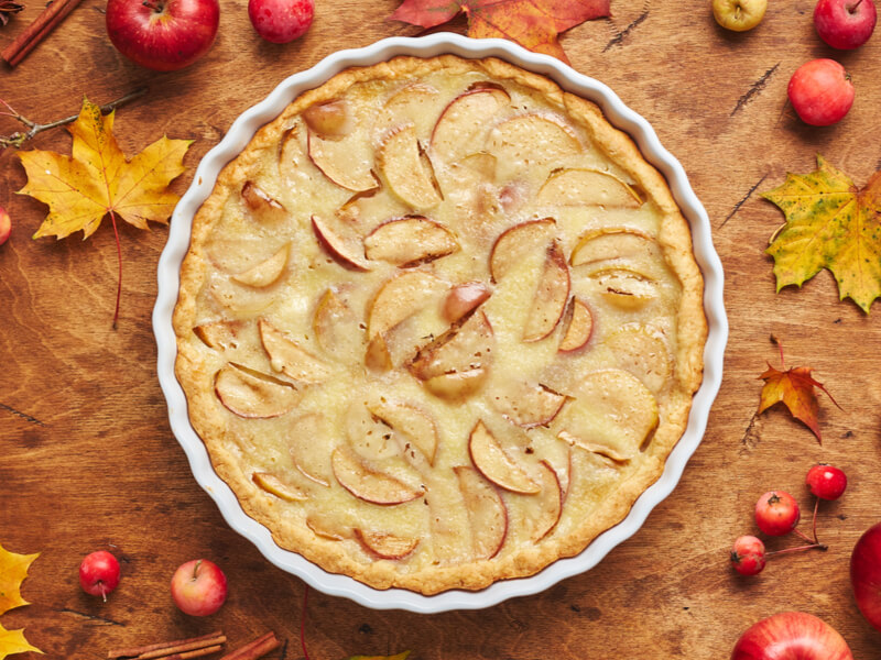 Torta de maçã em recipiente branco, sobre superfície marrom com frutas vermelhas e folhas em volta