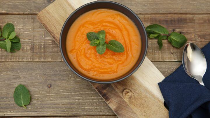 Sopa de cenoura em recipiente preto com hortelã em cima. Recipiente em sobre tábua