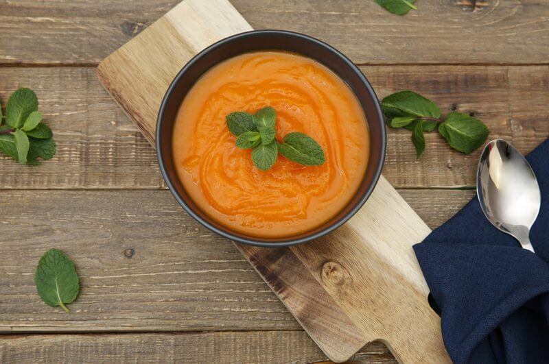 Sopa de cenoura em recipiente preto com hortelã em cima. Recipiente em sobre tábua