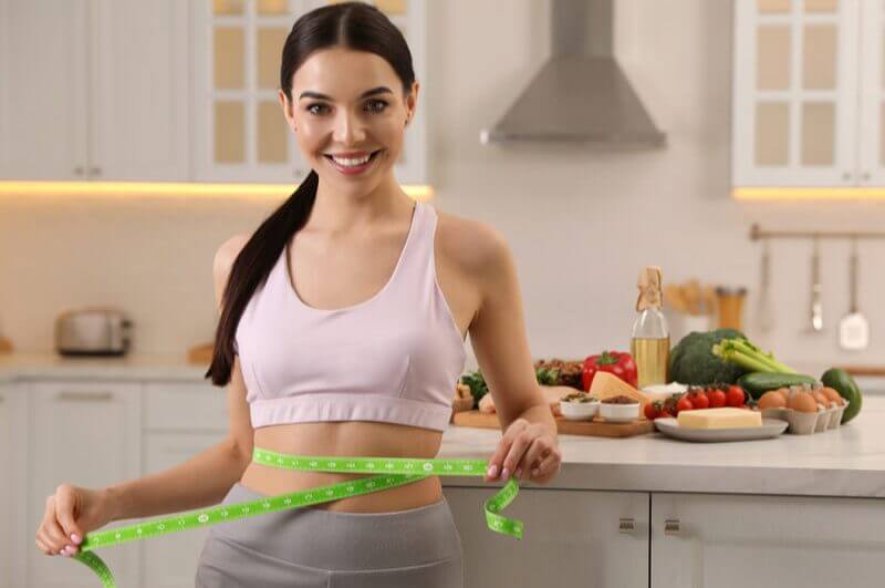 Mulher medindo a cintura com fita métrica e alguns alimentos saudáveis em cima da mesa