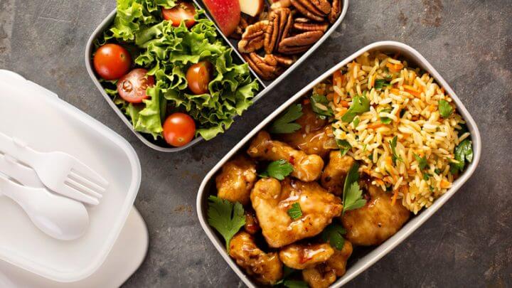 Duas marmitas cheias com arroz, frango, legumes e frutas e talheres de plástico ao lado