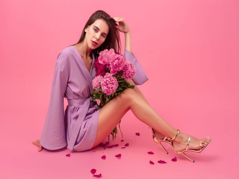 Mulher com vestido roxo e sapato dourado sentada em frente a um fundo rosa