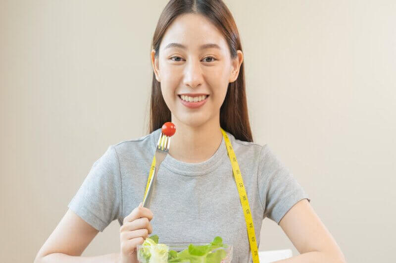 Mulher sentada sorrindo segurando um garfo com tomate e um recipiente com salada