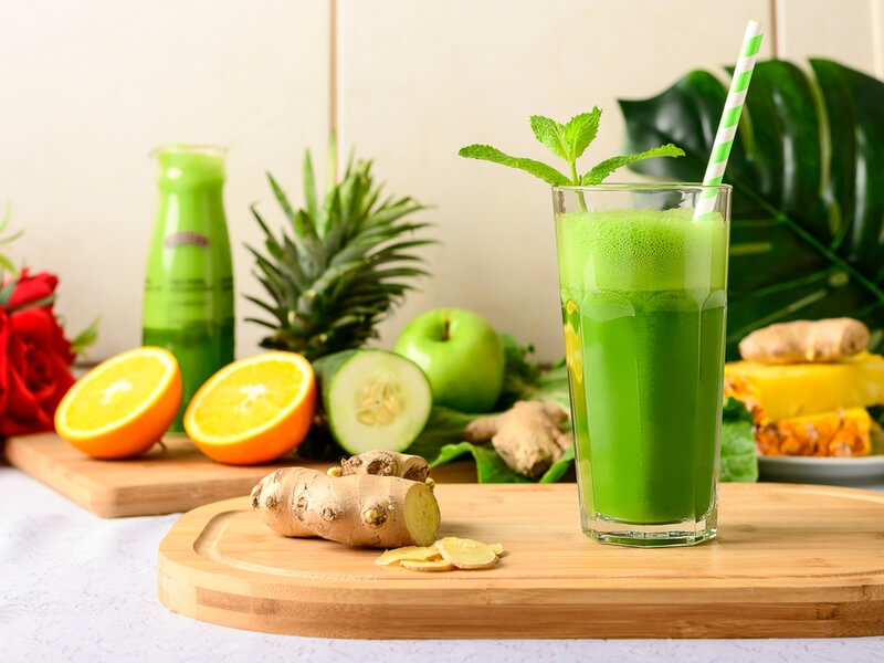 Copo com suco de couve e gengibre ao lado em cima da tábua de madeira e frutas ao fundo