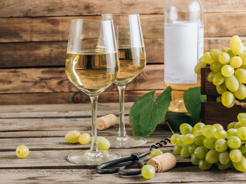 Duas taças com vinho branco, uvas verdes, saca-rolhas e garrafa de vinho ao fundo