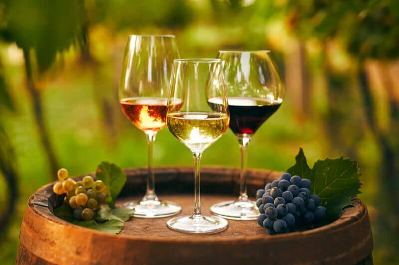 Três taças de vinho rose, branco e tinto em cima do barril e uvas ao lado