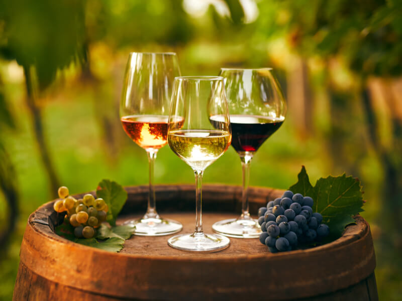 Três taças de vinho rose, branco e tinto em cima do barril e uvas ao lado