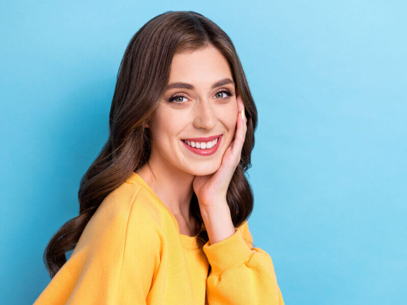 Mulher com cabelos longos e blusa amarela em frente a um fundo azul sorrindo com a mão no rosto