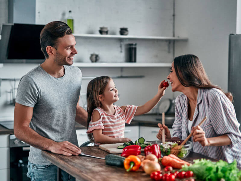 Pai, filha e mãe na cozinha preparando comida. Menina oferece um morango para a mãe