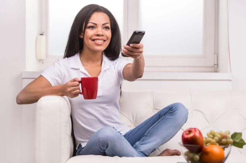 Mulher sorrindo em frente a TV, com o controle em uma mão e uma xícara em outra.