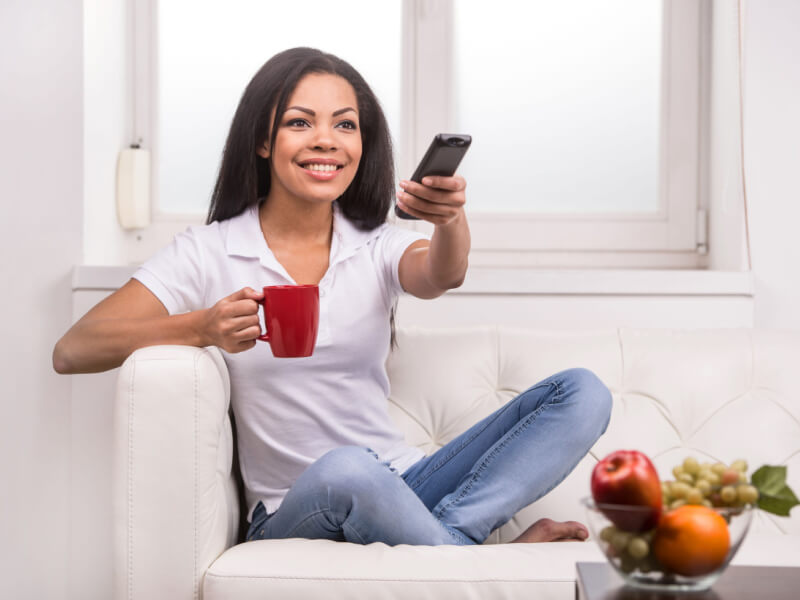 Mulher sorrindo em frente a TV, com o controle em uma mão e uma xícara em outra.