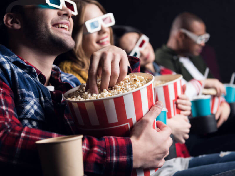 Jovens amigos sorridentes em óculos 3d comendo pipoca e assistindo filme no cinema