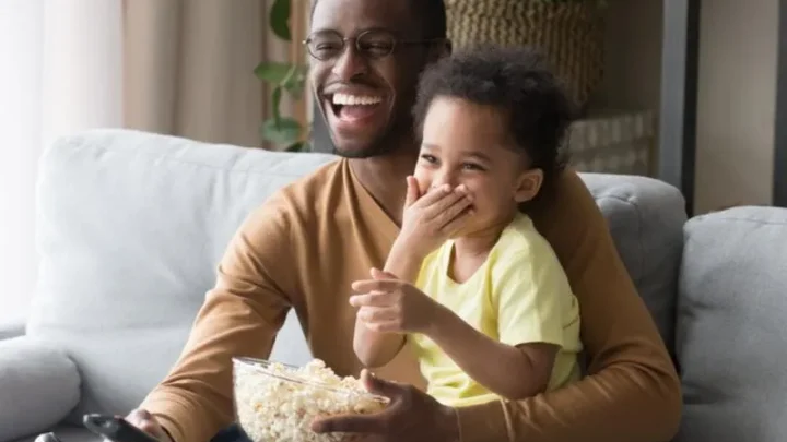 Pai e filho assistem a televisão sorridentes, e com pipoca.