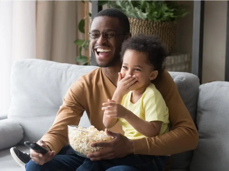 Pai e filho assistem a televisão sorridentes, e com pipoca.