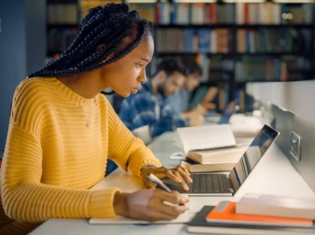 Na imagem, jovem estudante em uma biblioteca escrevendo no computador e em um caderno concentrada