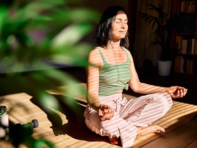 Mulher de regata verde e calça rosa meditando ao lado de uma planta