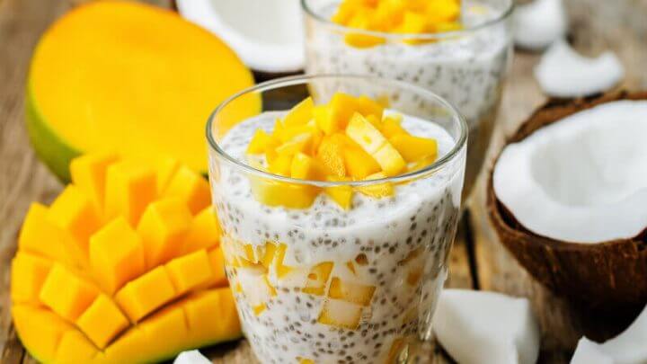 Copo de vidro com pudim de chia e pedaços de manga em cima de uma mesa de madeira