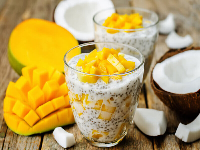 Copo de vidro com pudim de chia e pedaços de manga em cima de uma mesa de madeira