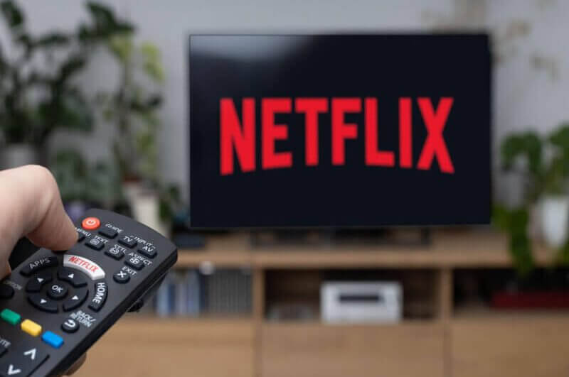 TV com logo da Netflix em rack marrom. Mão com controle remoto apontado para a TV