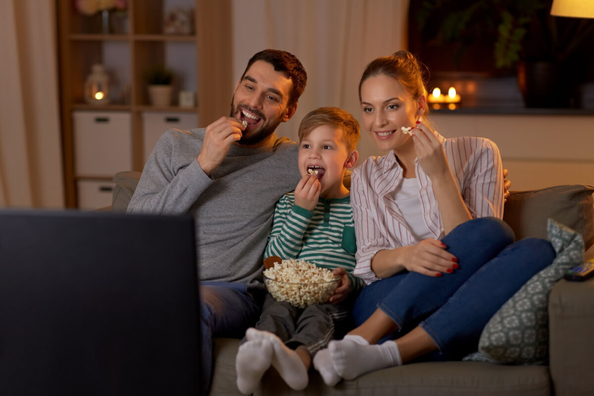 Pai, filho e mãe sentados no sofá vendo filme e comendo pipoca