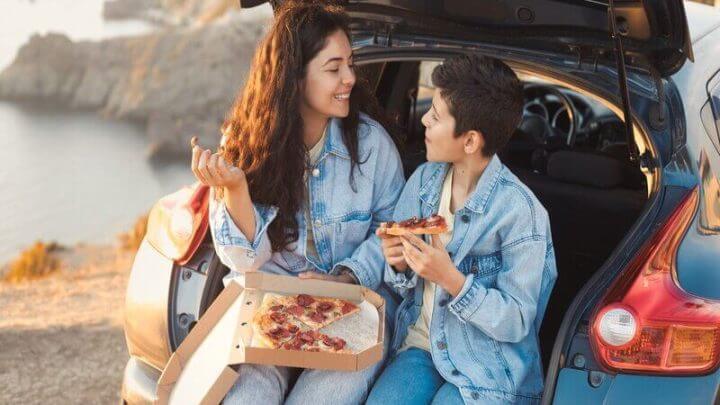 Mulher e menino sentados no porta malas de um carro comendo pizza