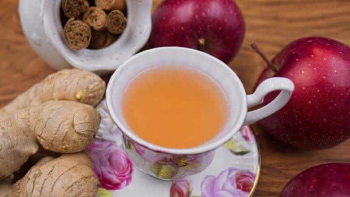 Uma xícara branca com detalhes em flores rosa cheia de chá termogênico; maças, gengibre e canela em pau estão ao seu redor