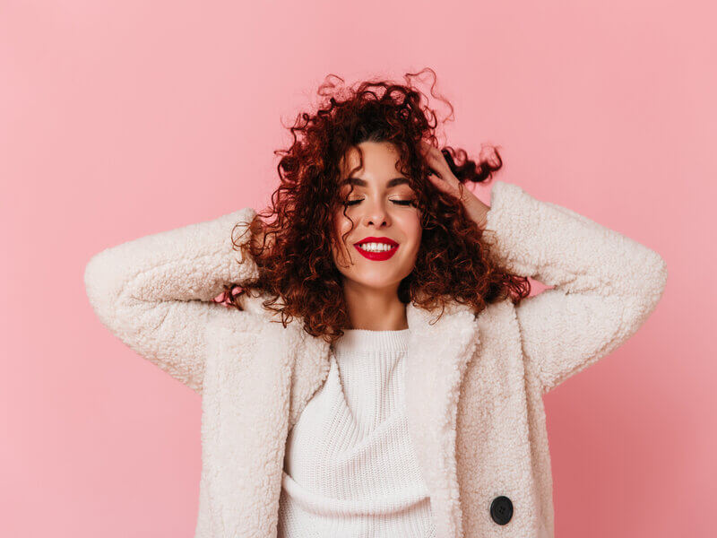 Mulher sorrindo com as mãos no cabelo em frente a um fundo rosa