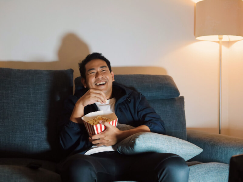 Homem alegre sentado no sofá e assistindo filme com pipoca.