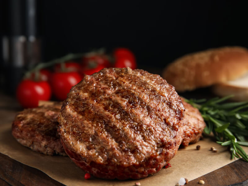 Hambúrguer de carne em cima de folha marrom. Tomates e alecrim ao fundo