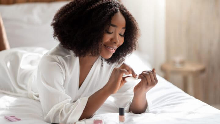 Mulher deitada na cama sorrindo e fazendo as unhas.