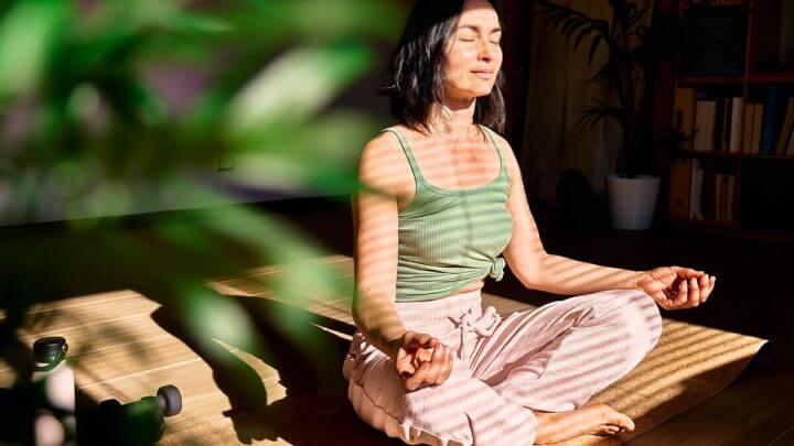 Mulher de regata verde e calça rosa meditando ao lado de uma planta.