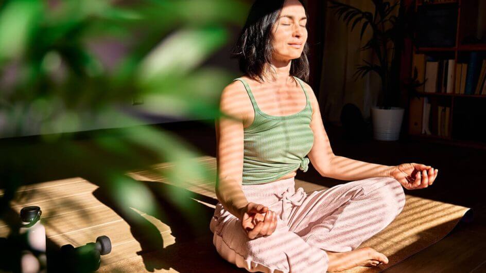 Mulher de regata verde e calça rosa meditando ao lado de uma planta.