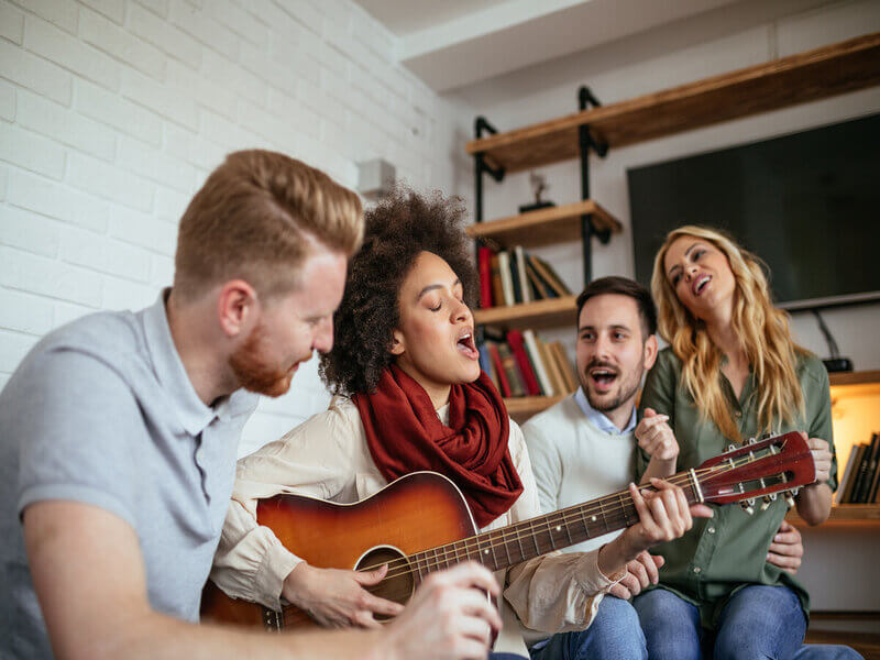Pessoas sentadas em sofá ouvindo uma mulher cantar e tocar violão