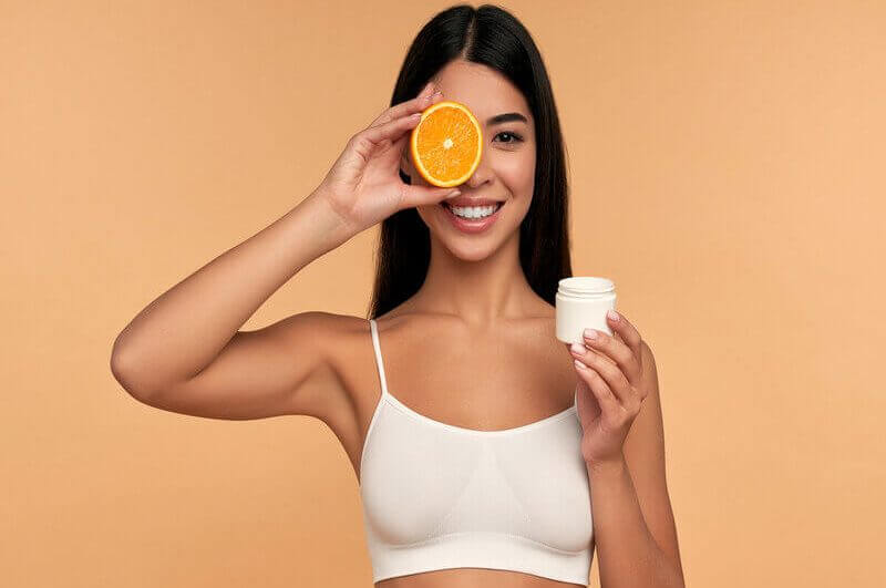 Mulher com top branco sorrindo segurando um recipiente branco e uma rodela de laranja em frente ao olho