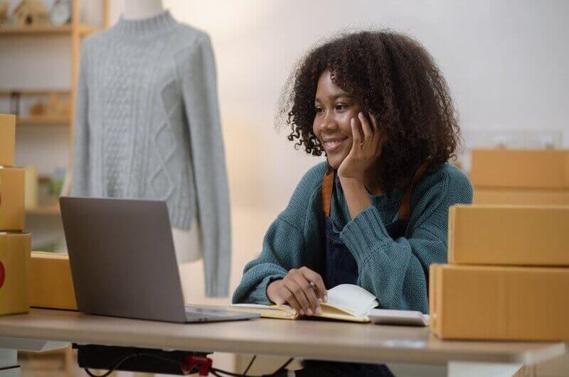 Mulher sentada sorrindo olhando para um notebook com caixas ao seu redor