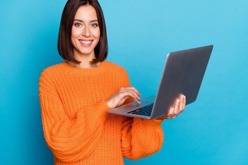 Mulher de blusa laranja sorrindo e segurando um notebook