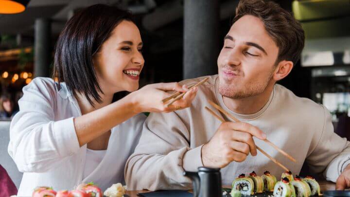 Casal comendo comida japonesa em uma mesa. Mulher oferecendo a ele comida