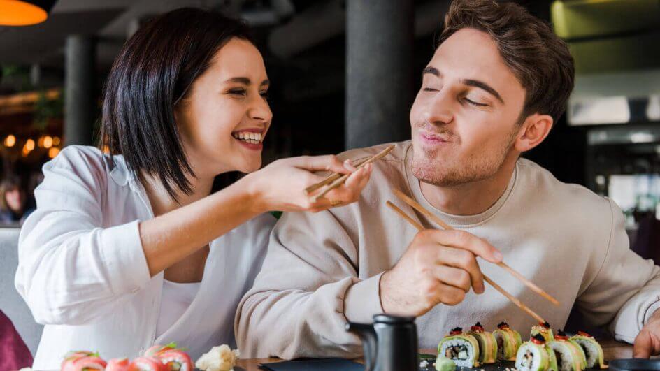 Casal comendo comida japonesa em uma mesa. Mulher oferecendo a ele comida