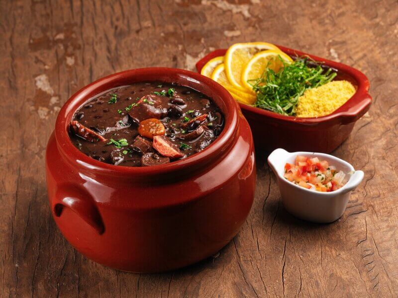 Recipiente de vidro marrom com feijoada e linguiça em cima de uma mesa de madeira com dois recipientes contendo laranja e tomate