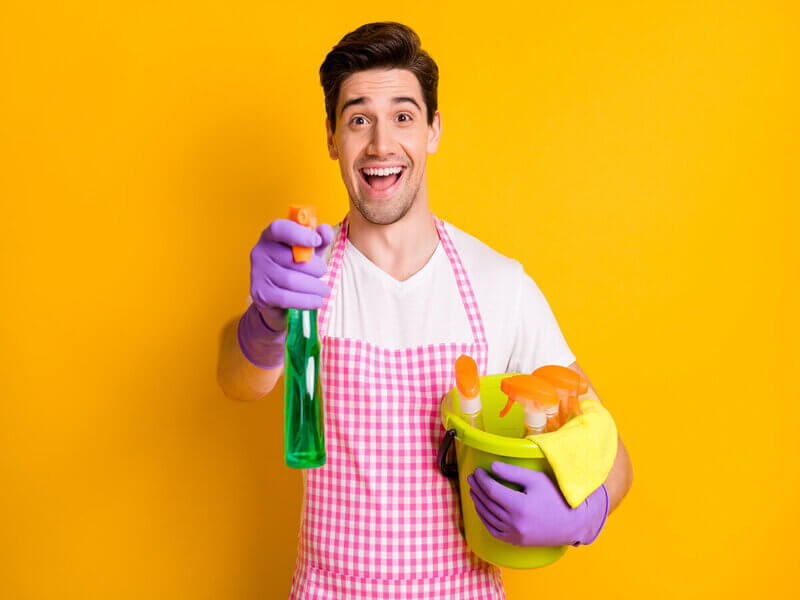 Homem com roupa de limpeza e luvas roxas sorrindo em frente a um fundo amarelo