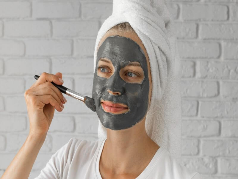 Mulher com toalha branca na cabeça, aplicando argila verde no rosto